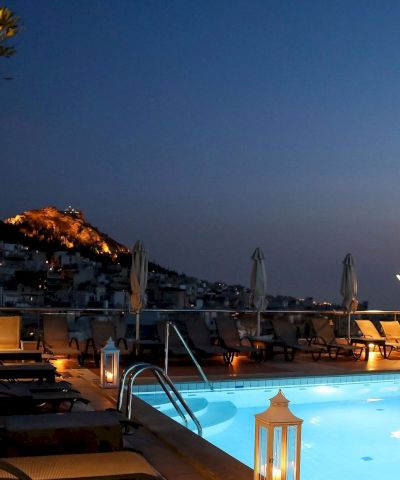 Rooftop swimming pool at dusk with lounge chairs and umbrellas, overlooking a hillside town lit up under a deep blue sky.