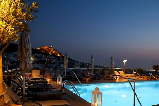 Rooftop swimming pool at dusk with lounge chairs and umbrellas, overlooking a hillside town lit up under a deep blue sky.