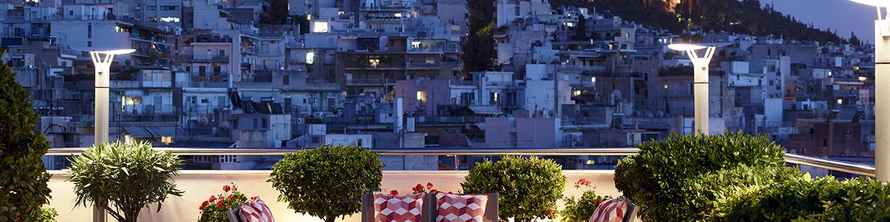 Rooftop terrace at dusk with floral planters and seating, overlooking a hillside town and a lit hilltop monument.
