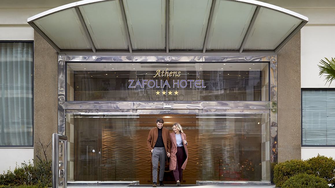 A hotel entrance with &ldquo;ZAFOLIA HOTEL&rdquo; signage and two people walking in front of the glass doors.