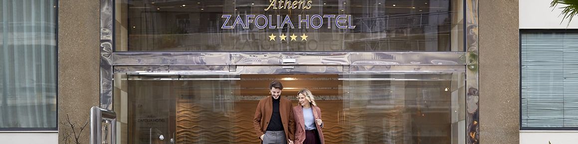 A hotel entrance with &ldquo;ZAFOLIA HOTEL&rdquo; signage and two people walking in front of the glass doors.