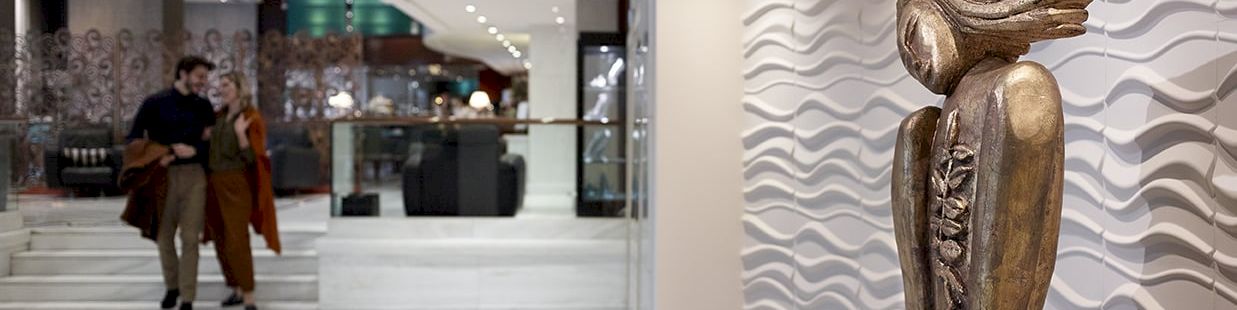 Modern lobby with shoppers and a bronze seated sculpture beside a wavy wall.