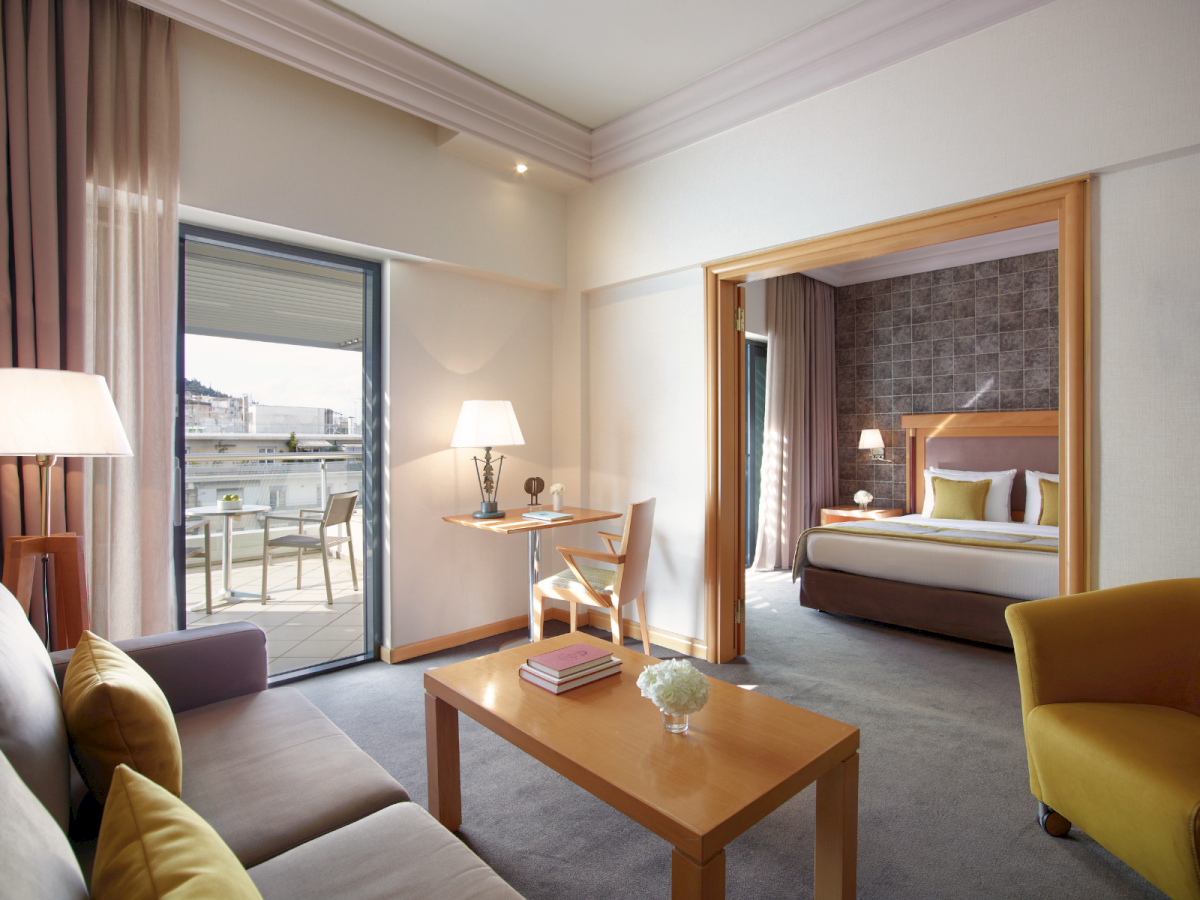 A modern hotel suite with sofas, a wooden coffee table, lamps, and an adjacent bedroom with a large bed and balcony view.