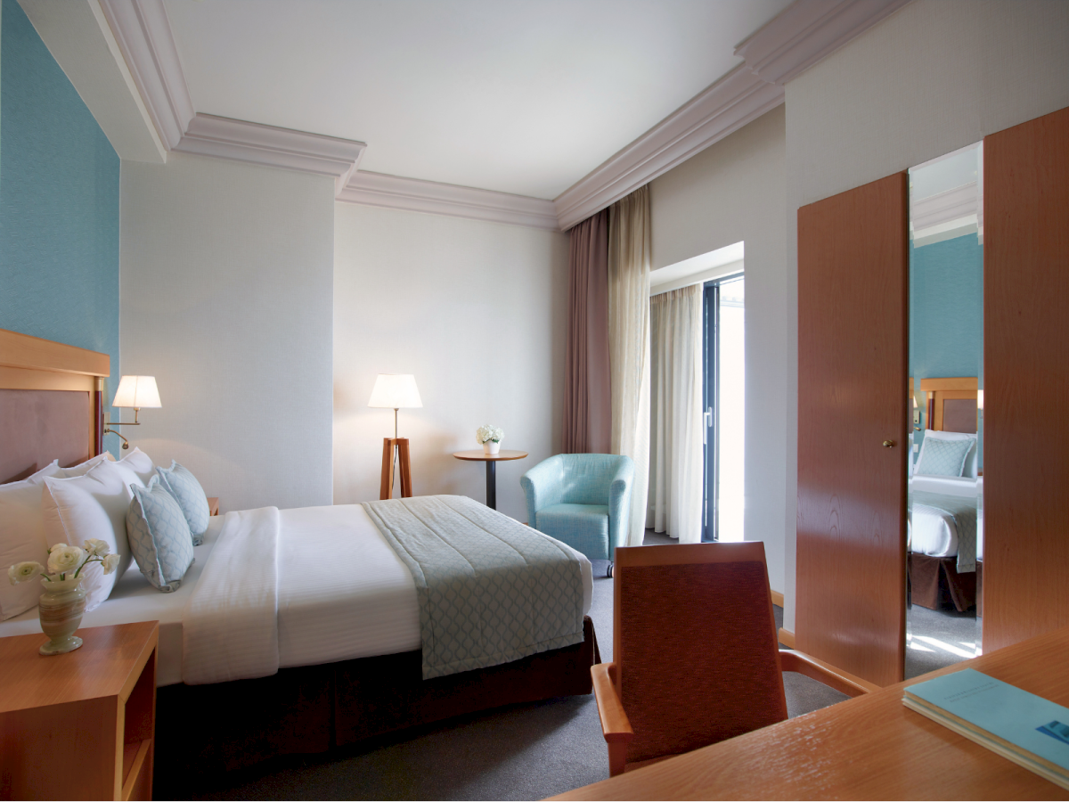 A bright hotel bedroom with a bed, bedside lamps, chairs, curtains, and an open door showing another room, ending the sentence.