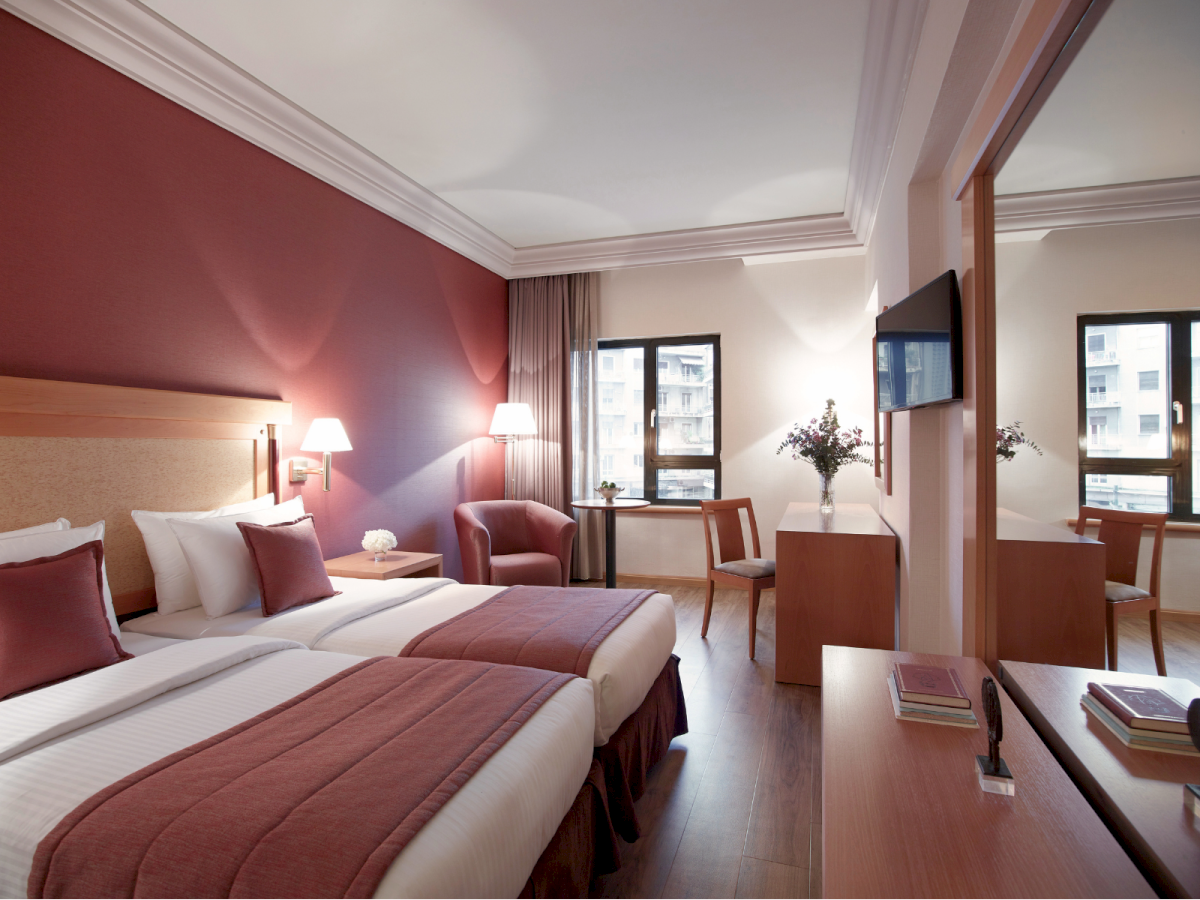 A modern hotel room with twin beds, pink curtains, a desk with books, and a wall-mounted TV.