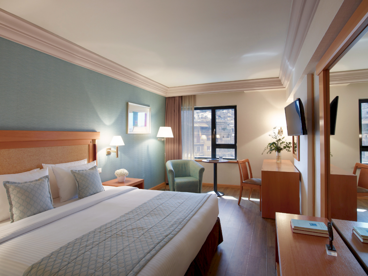 A hotel room with a neatly made bed, lamps, desk, two wall-mounted TVs, chairs, curtains, and a city view through the window.