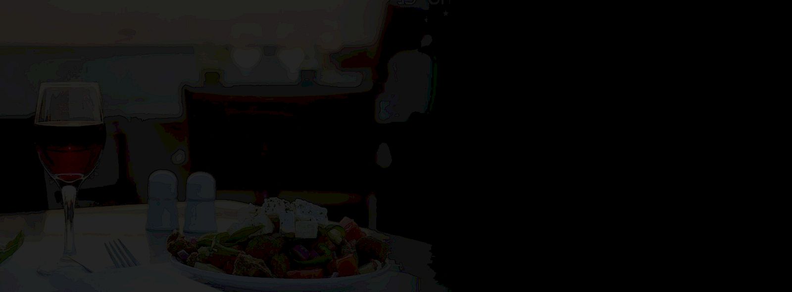 A glass of red wine sits beside a plated meal with salad and vegetables on a table in a dim dining room.