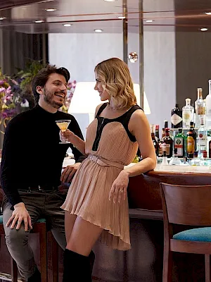 A couple chatting and smiling at a hotel bar, each holding a cocktail, with a row of liquor bottles behind them.