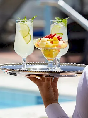 Two colorful cocktails with fruit and mint sit on a metal tray by a pool, ready to serve.