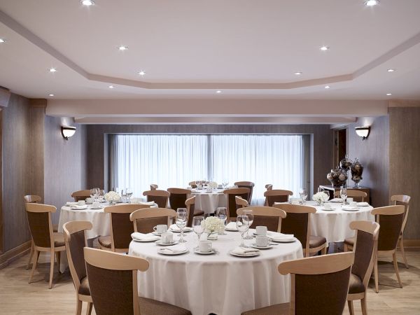 An upscale banquet room with round tables set with white linens, glassware, and chairs by tall windows.