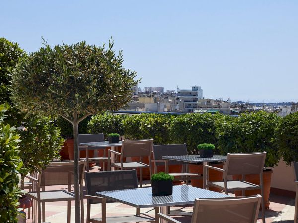 A sunny rooftop terrace with metal tables and chairs, surrounded by potted trees and hedges, overlooking a distant city skyline.