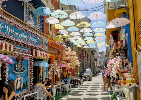 A sunny street lined with shops, packed with colorful umbrellas overhead and spring flowers, with a couple of people walking.