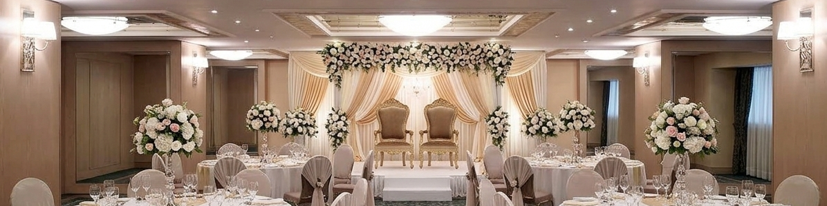 A beautifully decorated wedding reception hall with round tables, white chairs, and floral arches along the stage, inside.