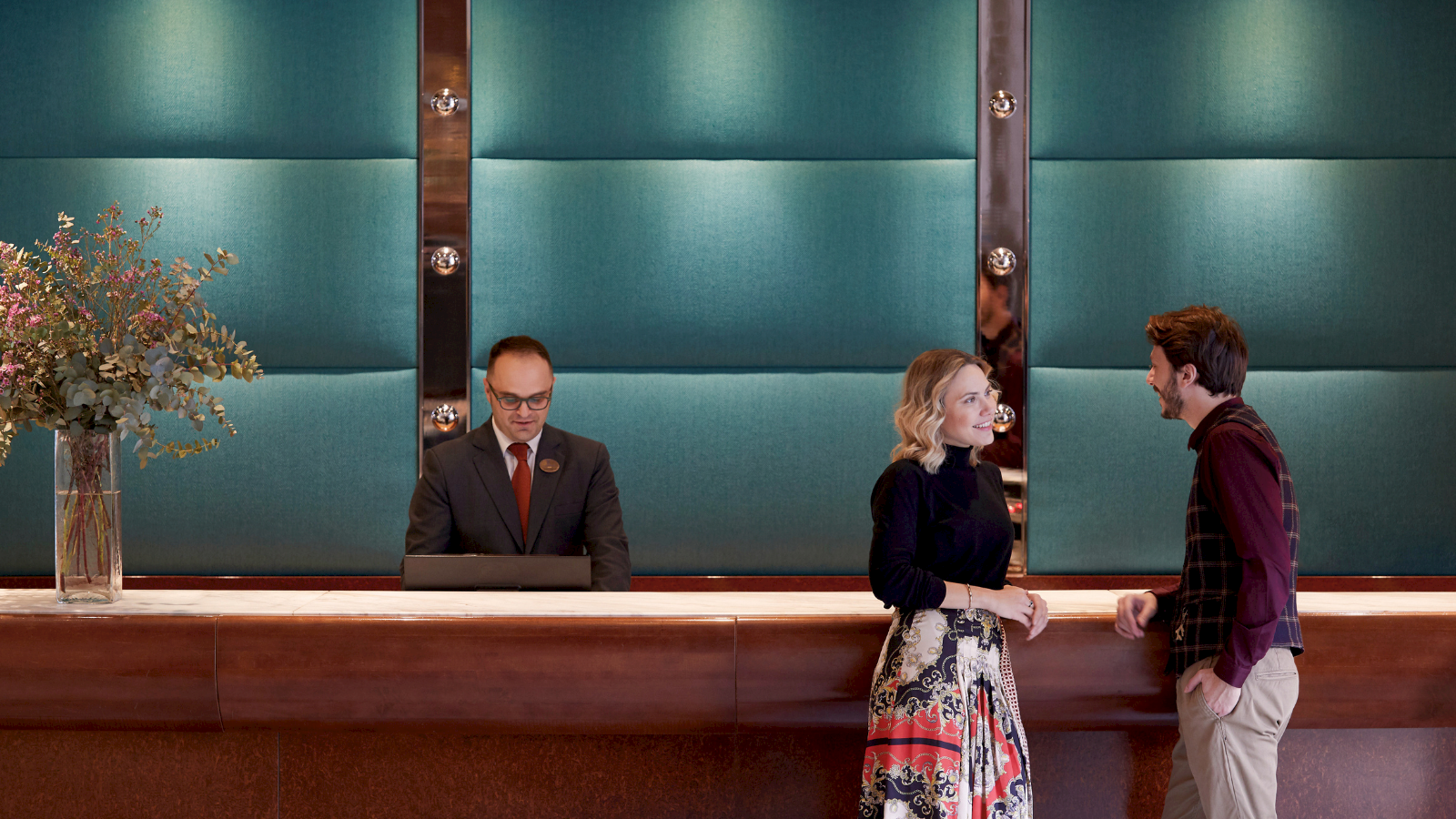 A man in a suit presents at a podium while two people chat in a modern conference lobby.