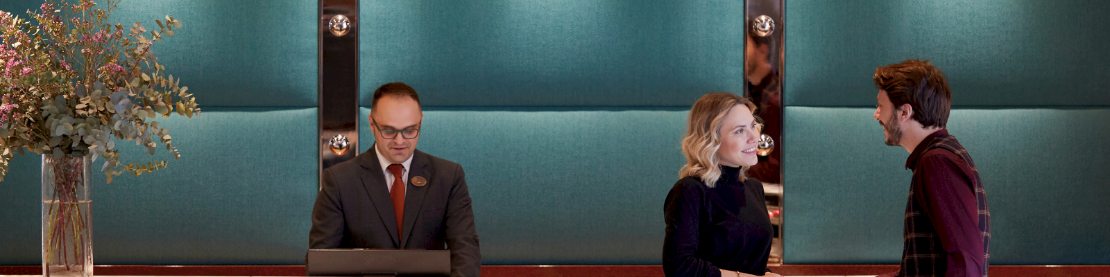 A man in a suit presents at a podium while two people chat in a modern conference lobby.