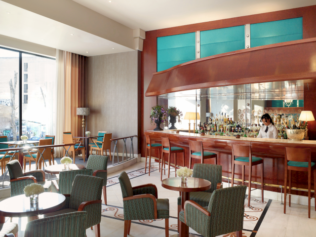 A modern hotel lounge with a bar counter stocked with bottles, green chairs, and a bartender serving guests.