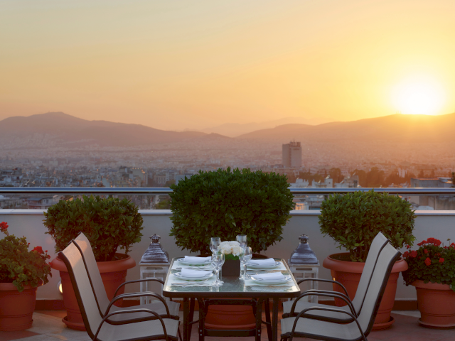 Outdoor terrace dining set with chairs, glasses, potted plants, and a golden sunset over a city and distant mountains.