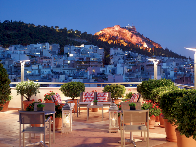 A rooftop terrace with potted flowers and chairs overlooks a nighttime city and a glowing mountain with a tower on top.