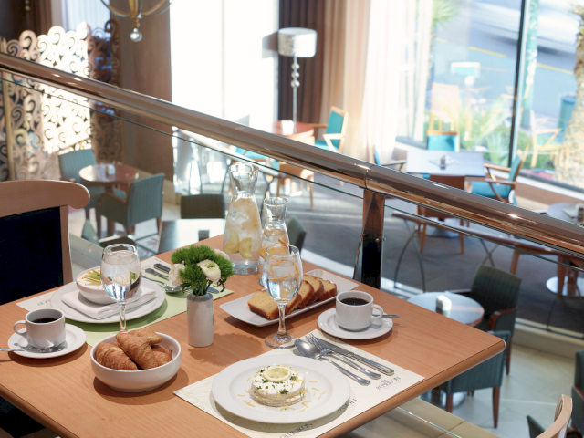 A sunny restaurant breakfast table with croissants, toast, a small cake, coffee and water, set by a big window view.
