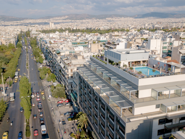Aerial view of a city boulevard next to a large modern hotel with balconies, rooftop pool, and parked cars.
