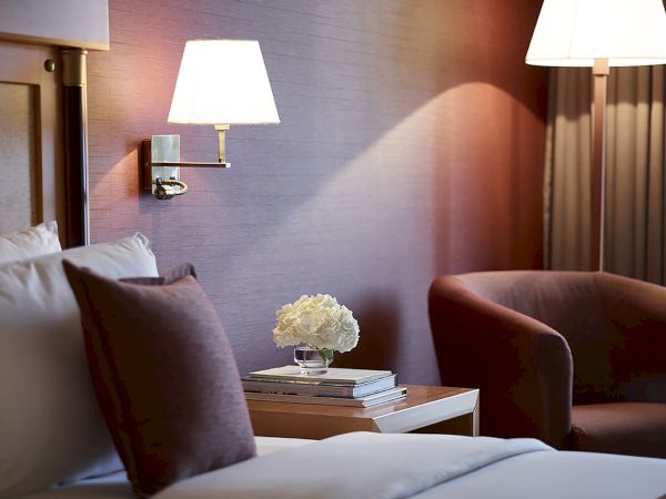 A cozy hotel room with a bed, armchairs, wall lights, and a small flower arrangement on a bedside table.
