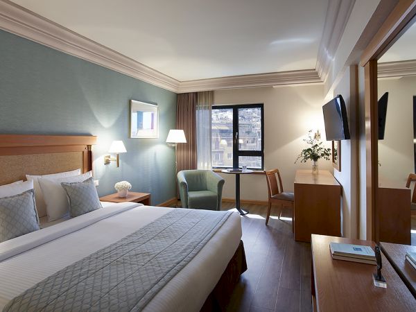 A modern hotel room with a neatly made bed, desk, chairs, lamps, TV screens, and large windows overlooking the city.