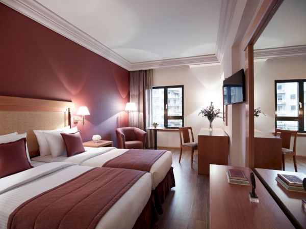 A modern hotel room with a double bed, lamps, armchairs, a desk, wall TV, and large windows with curtains.