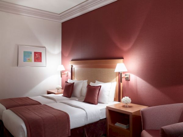 A styled hotel room with two beds, red-and-white pillows, bedside lamps, framed art, and a burgundy accent wall.