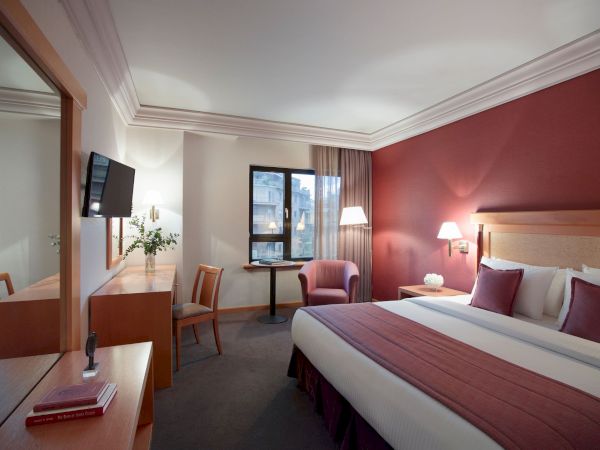 A modern hotel room with a king-size bed, desk, TV, armchair, lamps, and a window with curtains, all in warm colors.