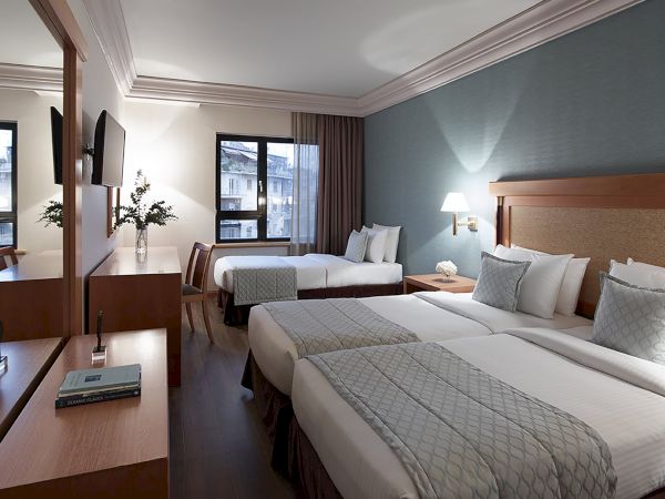 A modern hotel room with two twin beds, bedside lamps, curtains, and a view through large windows.