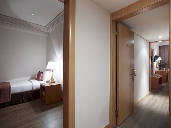 A modern hotel room with a bed, bedside lamp, and wooden doors leading to a hallway and dining area.