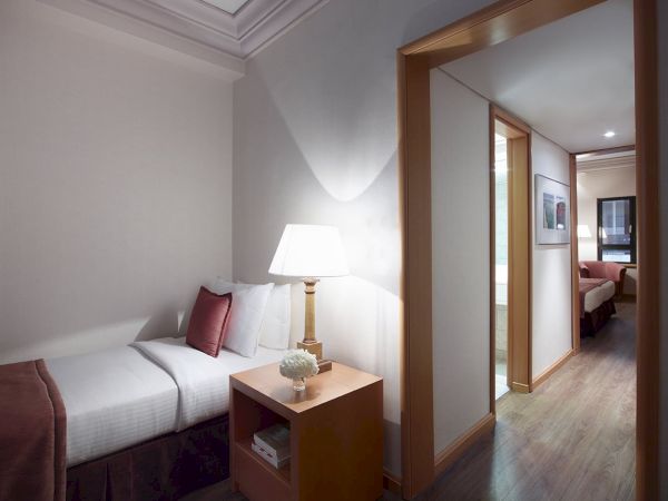 A modern hotel bedroom with a bed, bedside lamp, small table, and a doorway leading into a living area.