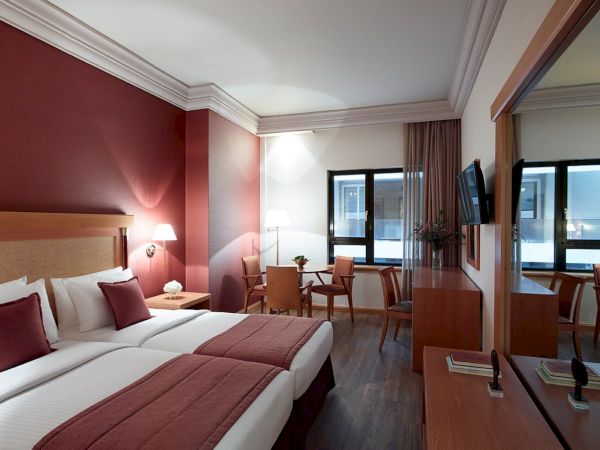 A modern hotel room with twin beds, chairs, a small desk, and large windows with curtains.