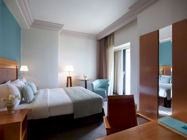 A modern hotel bedroom with a neatly made bed, bedside lamps, curtains, and an open door showing another room.