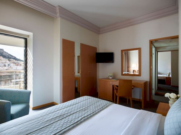 A modern hotel bedroom with a bed, desk, TV on the wall, wooden doors, and a window with city views.