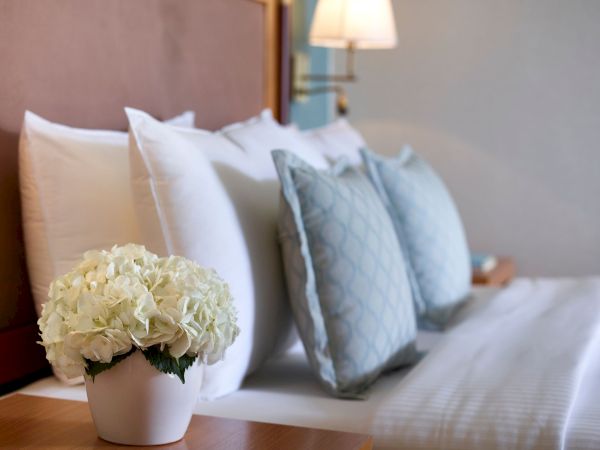 A bright, neatly made bed with white pillows and a blue patterned throw, topped by a vase of pale white hydrangeas.