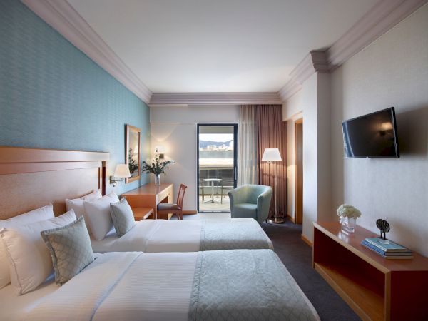 A modern hotel bedroom with beds, a lounge chair, desk lamps, a flat-screen TV, and curtains beside a balcony door.