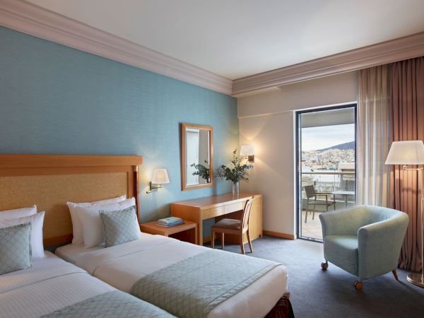A modern hotel bedroom with a double bed, bedside lamps, and a lounge chair by a large sliding window with city view.