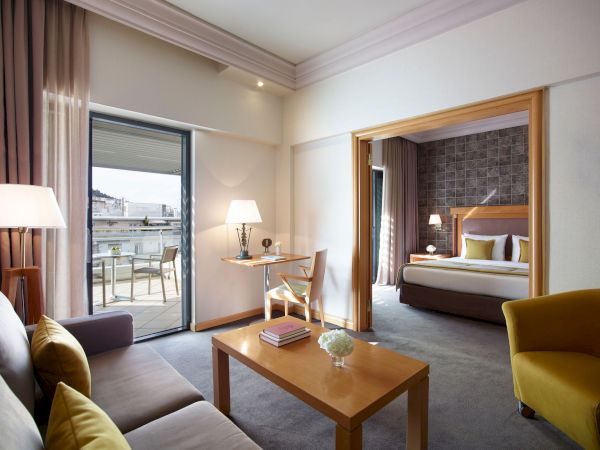 A modern hotel room with a sofa, coffee table, desk, lamps, and a bed, plus large doors opening to a balcony.