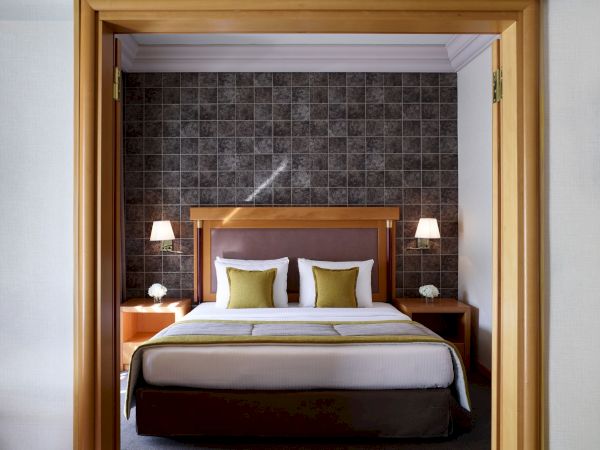 A hotel-style bedroom with a king-size bed, two lamps, and dark checkered wall panels seen through open wooden doors.