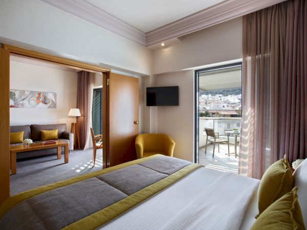 A modern hotel room with a bed, TV, sofa, floor lamp, and open balcony doors with city views, softly lit and neatly styled.