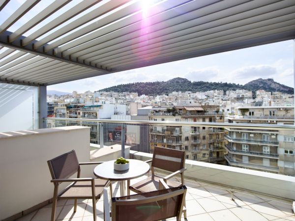 A sunny apartment balcony with table and chairs overlooks a dense cityscape and distant hills under a clear sky.