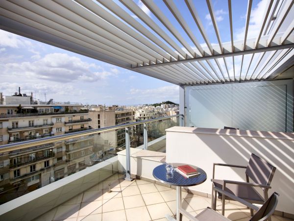A sunny apartment balcony with a round table, chairs, glass panels, and a striped metal canopy overlooking the city skyline.