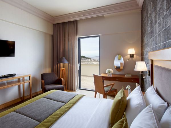 A hotel bedroom suite with a bed, desk, TV, lamps, curtains, and a balcony door overlooking the city.