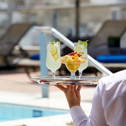 A server holds two colorful cocktails with fruit garnishes on a tray beside a sunny swimming pool.