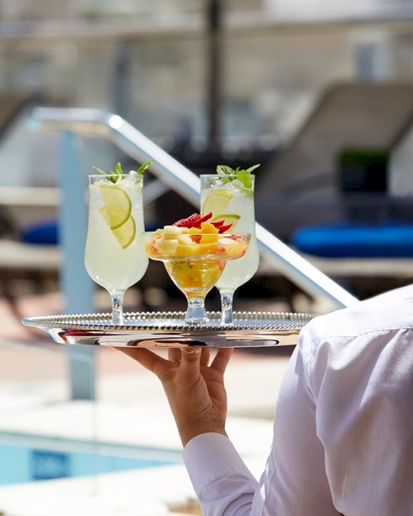A server holds two colorful cocktails with fruit garnishes on a tray beside a sunny swimming pool.