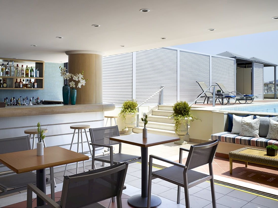 Rooftop lounge with a bar, seating, and a poolside area with sun loungers behind louvered windows.