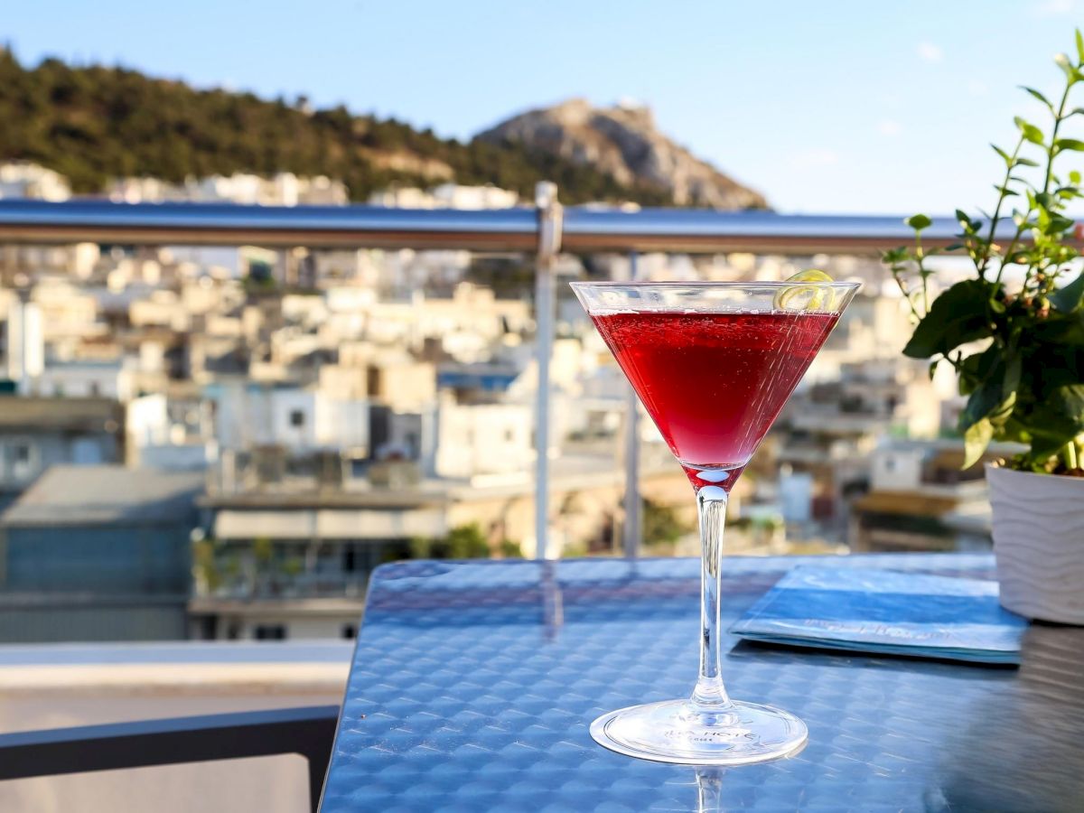 A red cocktail in a martini glass on a terrace table, with a seaside town and cliffs in the blurred background.