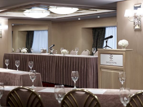 A hotel banquet hall with served tables, microphones, and a podium marked &ldquo;Athens ZAFOLIA HOTEL&rdquo;.