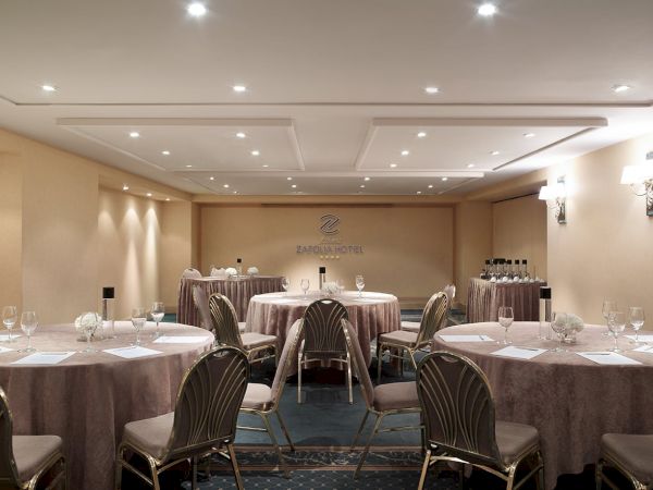 A hotel banquet room with round tables set for dinner, elegant chairs, and the Zafola Hotel logo at the back.